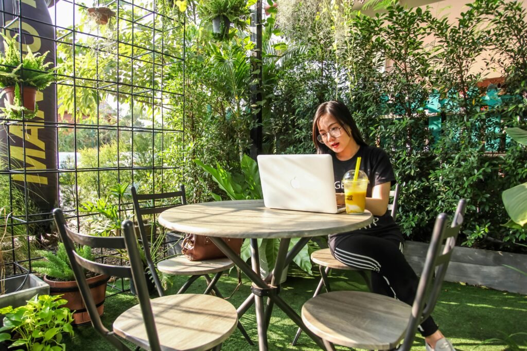 A woman working on a laptop in an outdoor garden setting, enjoying a refreshing drink.